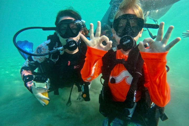Two scuba divers underwater making OK hand signs, wearing wetsuits and diving masks.