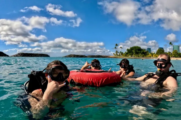 Four snorkelers in the ocean with a red float, tropical backdrop, and blue sky.