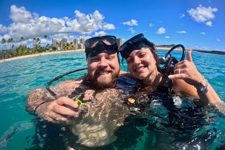 Two people in scuba gear smiling in clear water with a city skyline and palm trees in the background.
