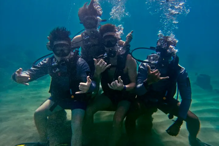 Four scuba divers posing underwater with hand signals and bubbles rising.