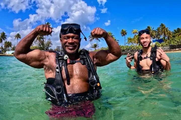 Two people in scuba gear smiling and posing in tropical beach water with palm trees in the background.