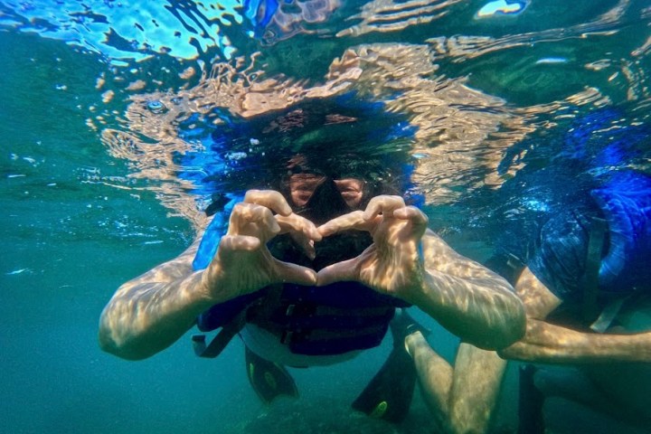Snorkeler underwater forming heart shape with hands, clear blue water.