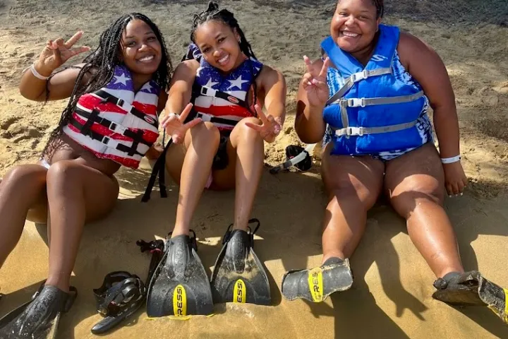 Three women in life vests sitting on sand, wearing snorkel fins, making peace signs.