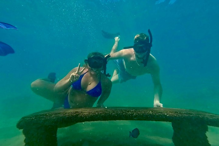 Two snorkelers underwater near a submerged bench, woman showing a peace sign.