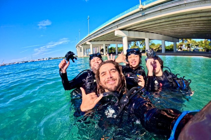 Four scuba divers posing for a selfie in clear blue water under a bridge.